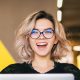 funny happy excited young pretty woman sitting at table in black shirt working on laptop in co-working office, wearing glasses